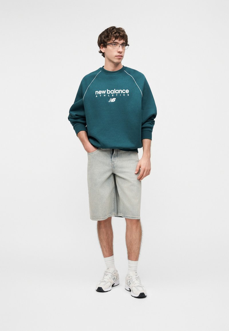 Jeune homme portant des lunettes, un sweat New Balance bleu sarcelle, un short en jean clair, des chaussettes blanches et des baskets blanches New Balance, debout les mains dans les poches.