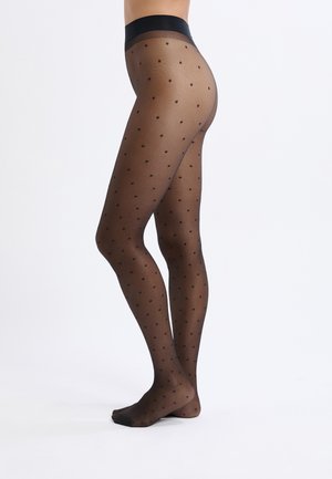 Mujer que lleva medias negras transparentes con un pequeño estampado de lunares, de pie descalza sobre una superficie blanca.