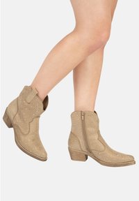 Botas de tobillo beige con acabado texturizado, adornadas con pequeños remaches, cierre de cremallera lateral y un bajo tacón apilado.