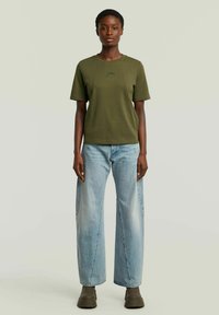 Vrouw staat rechtop met het gezicht naar voren, gekleed in een olijfgroen t-shirt, lichtblauwe wijde jeans en donkere bruine chunky laarzen.