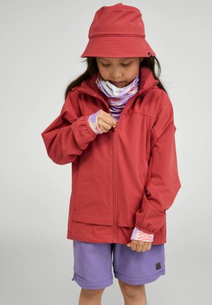 Child wearing red bucket hat and jacket, purple shorts, and patterned sleeves, zipping up jacket against neutral background.