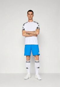 Bílé atletické tričko s černými pruhovanými akcenty, modré šortky, bílé ponožky s černými pruhy a bílé kopačky na fotbal.