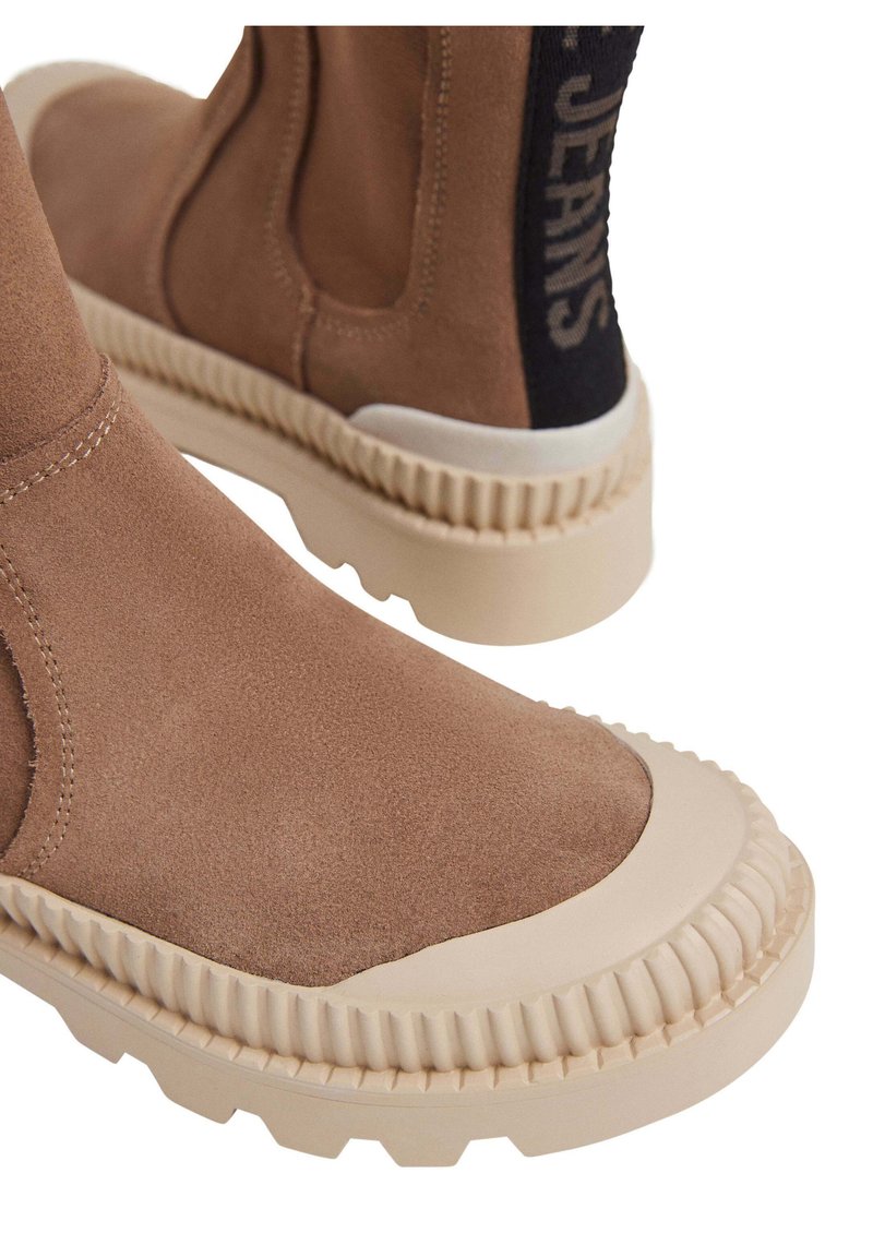 Brązowe zamszowe botki za kostkę z beżową gumową podeszwą, z teksturowanym brzegiem i czarną elastyczną wstawką oznaczoną napisem "JEANS" z tyłu.