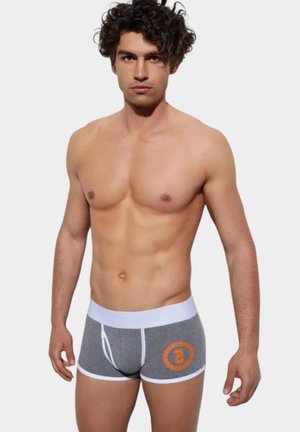 Młody mężczyzna o ciemnych, kręconych włosach, ubrany w szare bokserki z białą lamówką i pomarańczowym, okrągłym logo po lewej stronie, stojący na jednolitym tle.