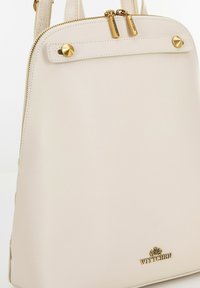 Beige leren rugzak met een gladde textuur, voorzien van gouden hardware, twee bovenste ritsen en decoratieve studs. Merkenlogo aan de onderkant.