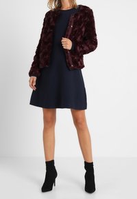 Bordowa kurtka z sztucznego futra o dopasowanym kroju, zestawiona z granatową sukienką w kształcie litery A. Czarne botki za kostkę dopełniają stylizację.