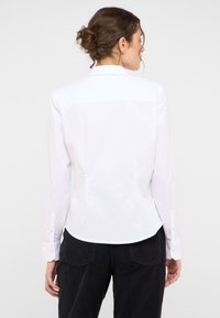 Biała koszula z długim rękawem, z kołnierzem odłożonym, dopasowana, z plisami z tyłu. Gładka faktura, mankiety na guziki i zaokrąglony dół.