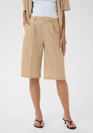 Persoon draagt beige knie-lange op maat gemaakte shorts met zijzakken, gecombineerd met een wit topje en zwarte sandalen met hak, staand op een witte achtergrond.