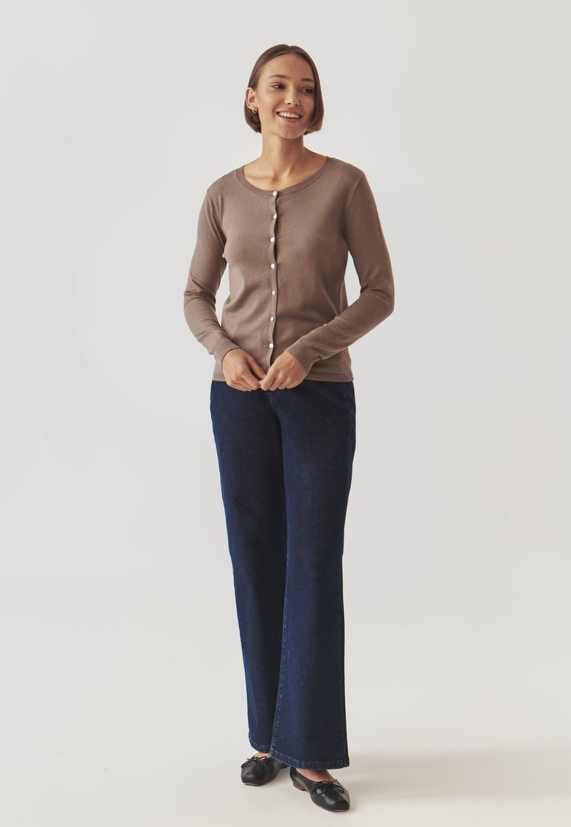 Bruine, lange mouwen cardigan met parelknopen, gecombineerd met donkerblauwe flared jeans. De stof lijkt soepel en de pasvorm is ontspannen.