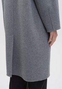 Grijze wolblendjas met een lange, iets oversized snit. Heeft een gladde textuur en een gebogen zoom, met een zichtbare buitenzak.