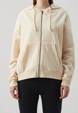 Persoon in een lichtbeige rits-hoodie met voorzakken en koordjes, gecombineerd met zwarte broek tegen een effen achtergrond.