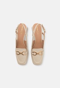 Světle béžové kožené boty na podpatku se čtveratou špičkou, zlatými detaily a polstrovanou stélkou. Obsahují nastavitelný pásek kolem kotníku.