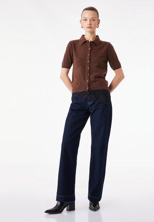 Kvinde iført en brun kortærmet knapcardigan, mørkeblå viddebenede jeans, sorte støvler med hæl, med håret trukket tilbage, stående mod hvid baggrund.