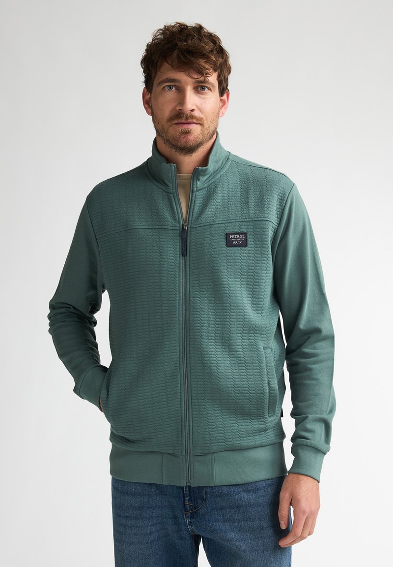 Chaqueta verde con cremallera, tela texturizada, cuello alto y parche con logo. Tiene puños y dobladillo acanalados. Se lleva sobre una camisa beige.