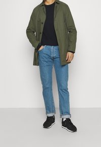 Olivgrön knapp-up kappa över en svart långärmad skjorta, ihop med ljusblå denimjeans och svarta träningsskor.