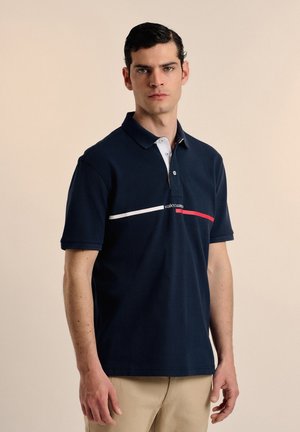 Polo de color navy hecho de algodón, con una franja horizontal blanca y roja en el pecho, y un cuello blanco con tres botones.
