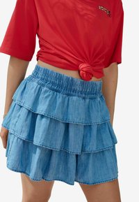 Jeansrock mit gestuftem Design und elastischem Bund, der drei Rüschenschichten in hellblau hat, präsentiert mit einem geknoteten roten T-Shirt darüber.