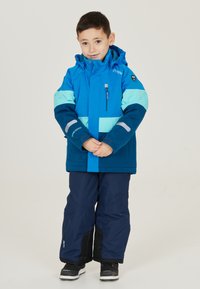 Blauwe en teal winterjas met capuchon, voorzien van een ritssluiting zak en reflecterende accenten. Gecombineerd met marineblauwe sneeuwbroek en zwarte laarzen.