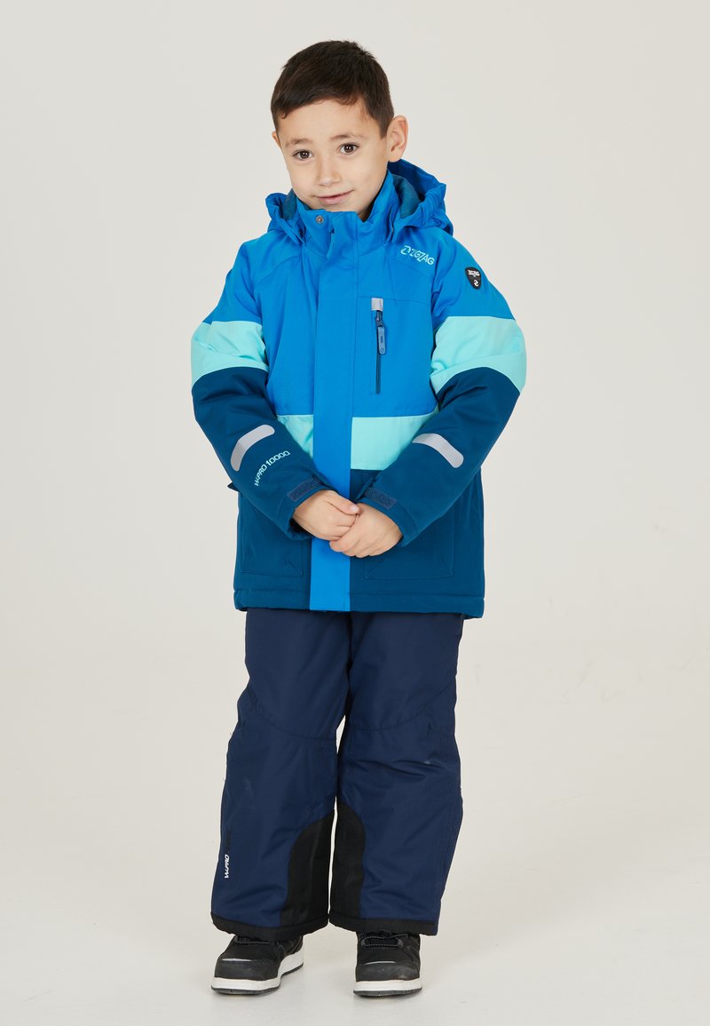 Blauwe en teal winterjas met capuchon, voorzien van een ritssluiting zak en reflecterende accenten. Gecombineerd met marineblauwe sneeuwbroek en zwarte laarzen.