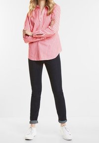 Chemise boutonnée à rayures rouges et blanches avec manches longues, associée à un jean foncé et des baskets blanches. La chemise présente un col et une coupe décontractée.