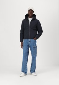 Chaqueta acolchada de color azul oscuro con capucha, cierre de cremallera y puños elásticos, combinada con jeans cargo de color azul claro y zapatillas blancas. Acento de gorra blanca.
