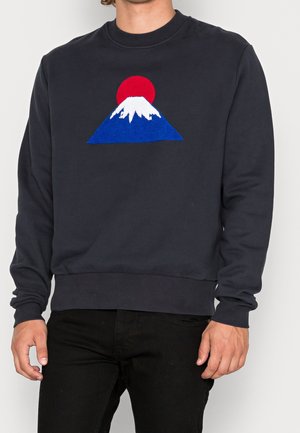 Mann trägt schwarzen Sweatshirt mit Grafik eines blauen Berges, bedeckt mit weißem Schnee und roter Sonne darüber, kombiniert mit schwarzer Hose.