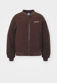 Tmavě hnědá bomber bunda New Balance se zipem vpředu, žebrovaným límcem, manžetami a spodním lemem, s malým bílým logem na hrudi.