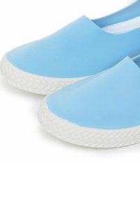 Zapatillas ligeras de color azul claro, sin cordones, con una parte superior suave y suela de goma blanca texturizada que presenta un patrón trenzado. Diseño de puntera redondeada, con detalles mínimos.