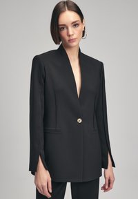 Blazer negro con escote en V profundo, hombros estructurados, cierre de un solo botón dorado y aberturas laterales en las mangas. Textura de tela suave.