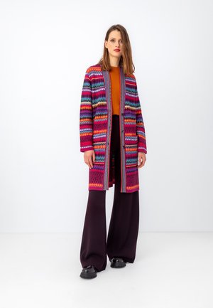 Multicolored knitted cardigan with geometric patterns, open front, and long sleeves, paired with a mustard top and wide black pants.