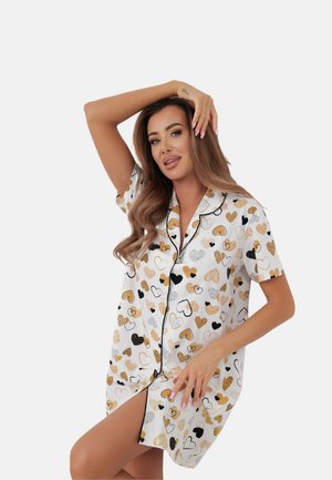 Kobieta w białej jedwabnej koszuli nocnej z beżowo-czarnymi i białymi wzorami serc, pozująca z podniesionym ramieniem i dłonią na głowie.