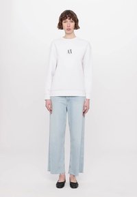 Biele crewneck mikina s grafickým logom, skombinovaná so svetlomodrými širokými nohavicami a čiernymi balerínami. Jednoduchý, neformálny outfit na neutrálnom pozadí.