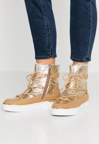 Sneaker alte metallic oro e sabbia con finitura in suede, dotate di una cerniera laterale e design con lacci, posizionate su una suola bianca.