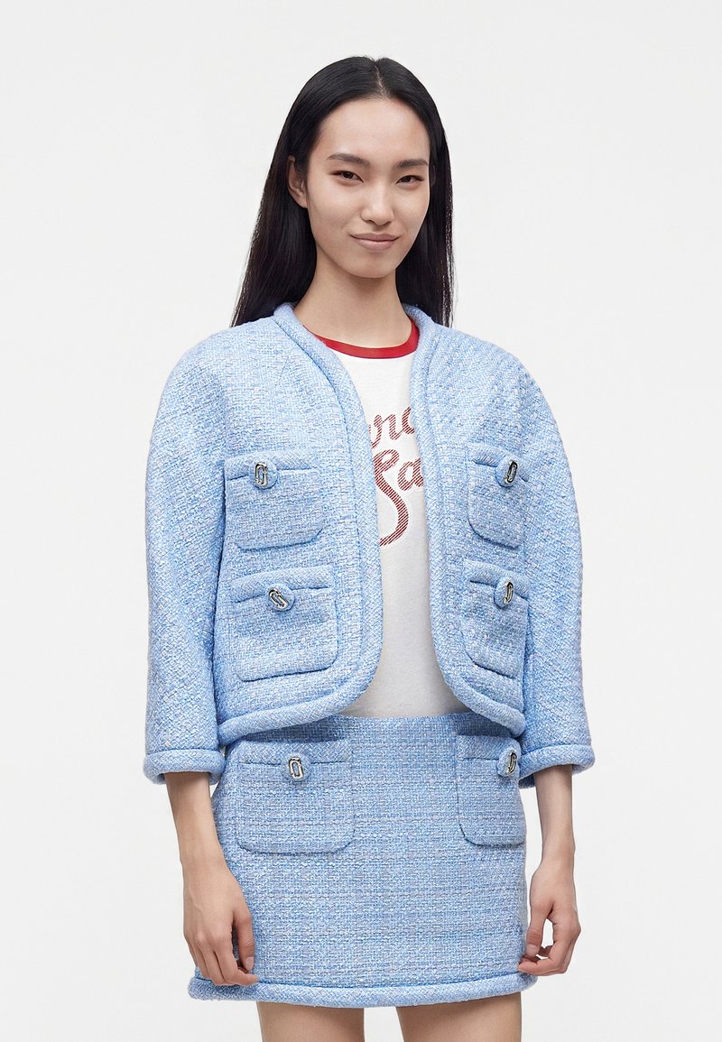 Femme aux longs cheveux noirs portant une veste texturée bleu clair et une jupe assortie avec quatre poches à l'avant, sur un t-shirt blanc à motif graphique.