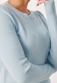 Persoon draagt een lichtblauwe trui met lange mouwen, met de ene arm gekruist en de andere hand bij het gezicht, tegen een witte achtergrond.