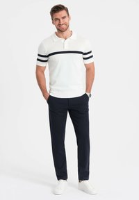 Witte polo-shirt met marineblauwe strepen, korte mouwen, ribgebreide kraag; gecombineerd met marineblauwe broek en witte sneakers. Model staat met handen in de zakken.