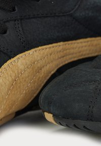 Gros plan sur des baskets en daim noir avec une bande en cuir marron texturé et des coutures détaillées sur la tige et les bords de la semelle.