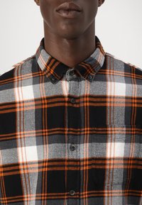 Zwart-witte geblokte shirt met oranje accenten, met een geknoopte kraag en een opgestikte zak, gemaakt van gestructureerde stof.