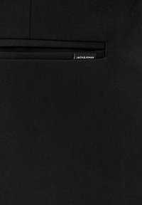 Gros plan sur un tissu noir avec une poche fendue horizontale, ornée d'une petite étiquette de la marque Jack & Jones en noir et blanc.