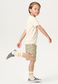 Kind trägt ein hellbeiges T-Shirt, olivgrüne Shorts und schwarz-weiße karierte Slipper mit creme- und gelb gestreiften Socken.