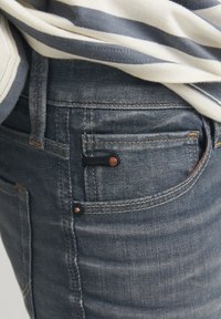 Tamno plave traperice imaju izbliza prikaz stražnjeg džepa s kontrastnim šavom i ukrasnom kopčom.