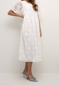 Valkoinen pitsimekko, jossa on lyhyet hihat, vartalonmyötäinen yläosa ja levenevä hameosa. Kangas on koristeltu pyöreillä kukkakuoseilla. Yhdistetty sinisiin korkokenkiin.