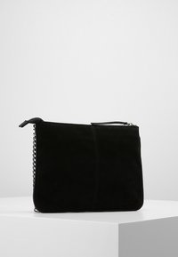 Bolso de mano de ante negro con cremallera y detalle de cadena plateada, exhibido en un pedestal blanco contra un fondo gris.