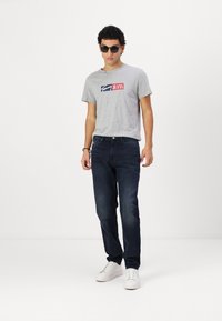 Lys grå kortermet t-skjorte med "Tommy Jeans" logo, kombinert med mørkeblå jeans og hvite sneakers. Modellen har på seg stilige svarte solbriller.