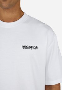 Valge puuvillane T-särk, millel on must tikand "PEGADOR Whispers of Gold" vasakul rinnal. Lihtne ümmargune kaelusdisain.