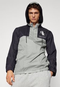 Lightweight, half-zip jacket featuring a gray body with black sleeves, adjustable hood, and logo detailing on the chest. Smooth texture.
