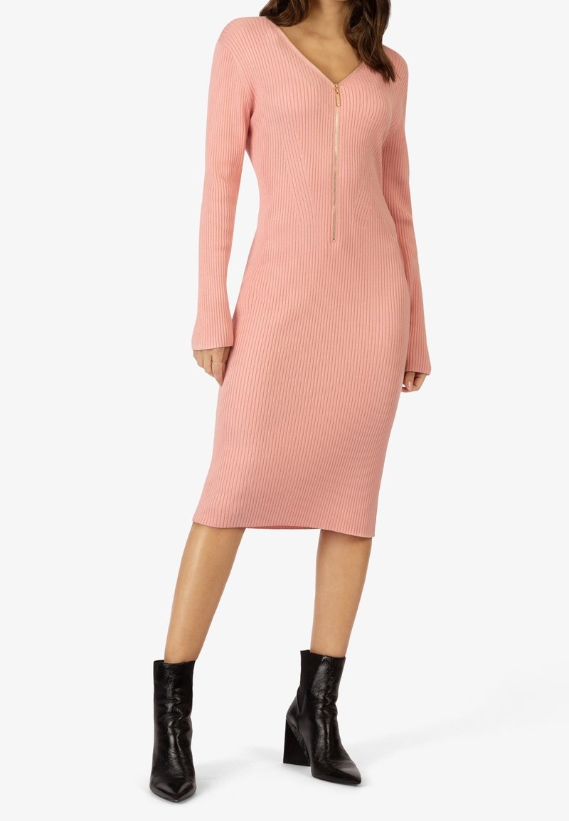 Femme portant une robe midi rose côtelée à manches longues avec une fermeture éclair avant, associée à des bottines à talons noires.