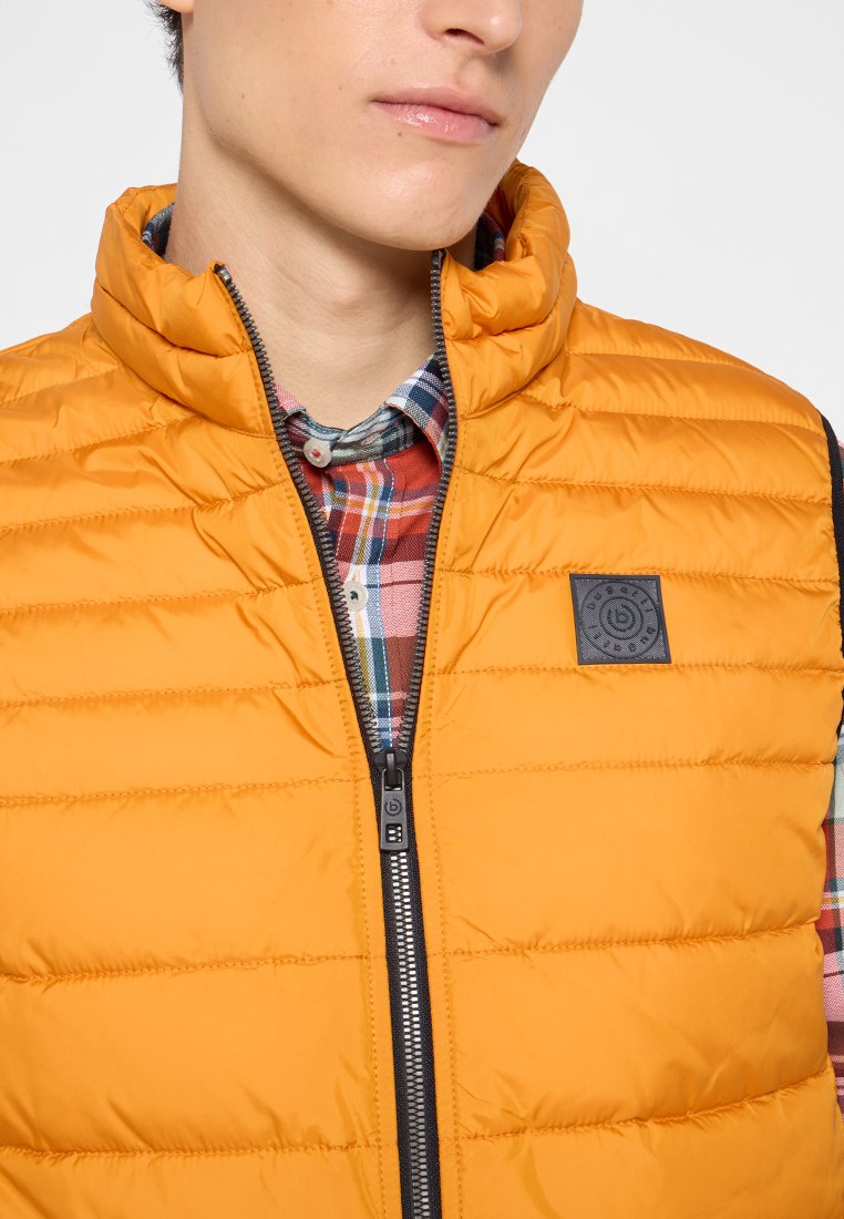 Gilet rembourré orange avec un col montant, fermeture éclair et écusson noir. En dessous, une chemise à carreaux aux tons rouges, verts et bleus est visible.