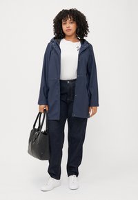 A person with curly hair wears a navy raincoat over a white shirt and dark jeans, holding a black handbag, standing against a neutral background.