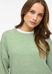 Gaiši zaļš adīts džemperis ar apaļu kakla izgriezumu un noslīpētām pleciem. Mīksta tekstūra, ar baltu apakškrekliņu.